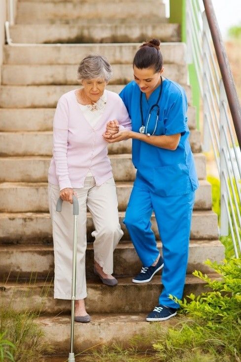 Caregiver in blue scrubs helping senior woman wit cane down some stairs outside | Senior Transportation and Errand Services | Neighborly Home Care