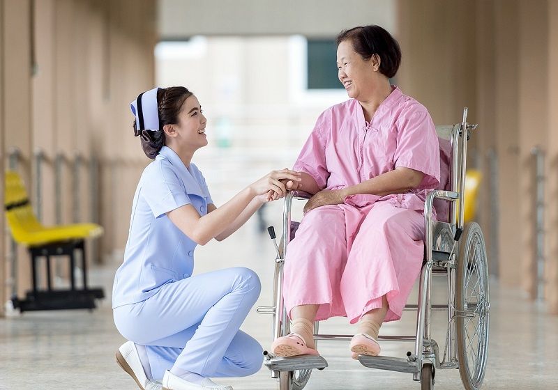 Caregiver kneeling beside woman patient in wheelchair in a hospital | In Hospital Private Duty Care  | Neighborly Home Care