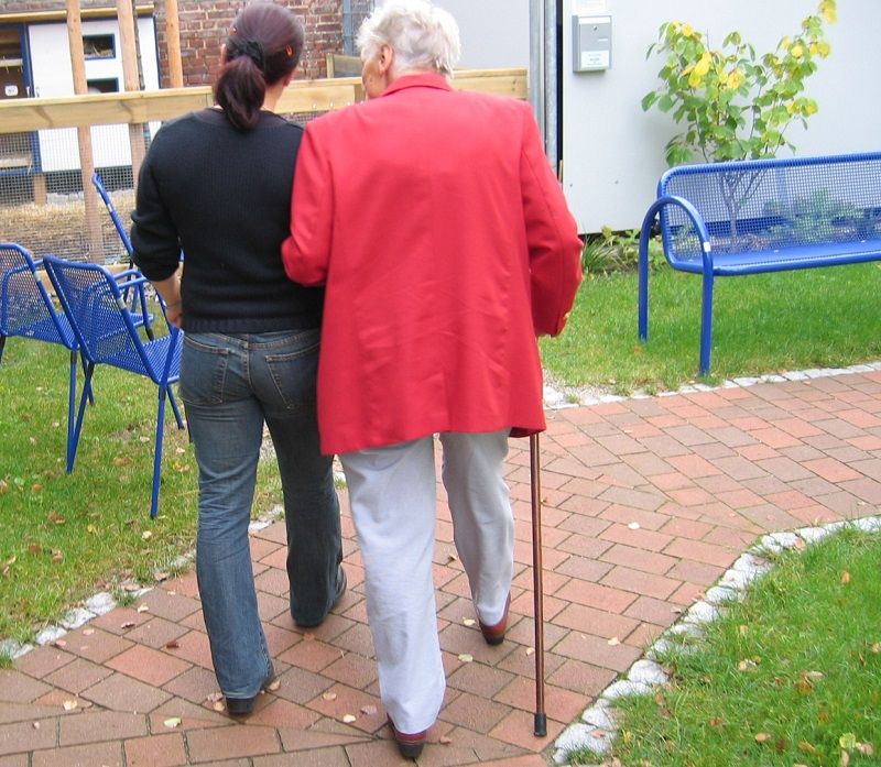 Younger woman walking with older woman who is using a cane down a path facing away from the camera | Senior Transportation Services | Neighborly Home Care