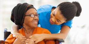 Caregiver leaning over the shoulder smiling and holding the hands of an elderly woman who is also smiling - Addressing the needs of the elderly - Neighborly Home Care